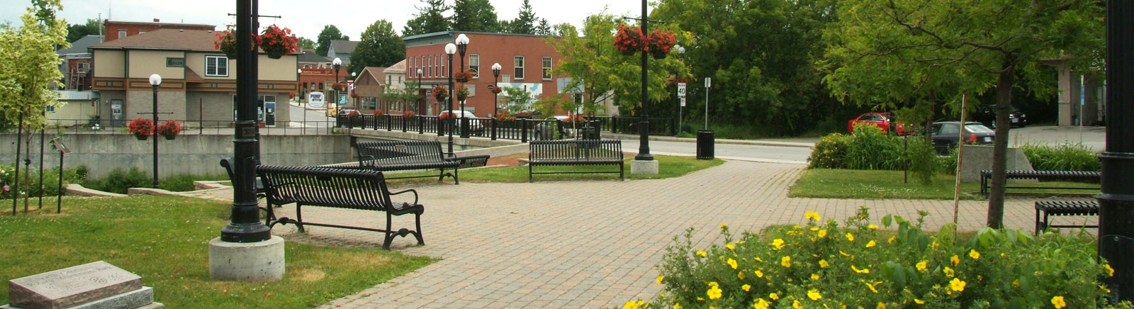 A view of a paved Kemptville park with benches and older buildings in the summer.